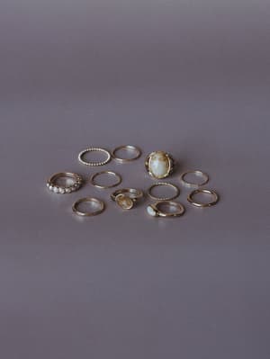 A scattered collection of gold stacking rings arranged casually on a muted gray surface — mixing dainty bands, pearl accents, twisted rope details, and statement stone rings in creamy ivory and warm amber tones.