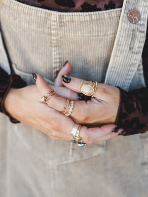 A close-up of layered hands showing off a curated stack of gold rings — stone, pearl, and delicate bands — against the texture of a sandy corduroy overall and a dark floral sleeve peeking through.