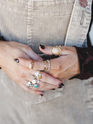 A close-up of layered hands stacked with an eclectic mix of gold, silver, and stone rings — oval cabochons, pearl clusters, turquoise accents, and crystal florals — worn against a corduroy overall and a dark floral sleeve, with deep plum nails tying