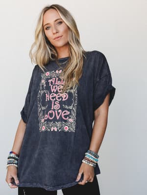 An oversized acid-wash charcoal graphic tee with a floral 'All We Need Is Love' print, styled with stacked beaded bracelets and layered gold necklaces — the kind of effortless, expressive look that feels just as good as it looks.