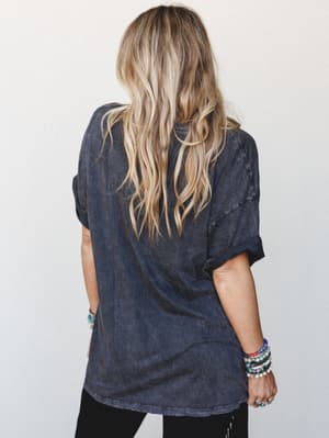 A back-facing shot of a model in an oversized acid-wash charcoal graphic tee with rolled sleeves, styled with stacked beaded bracelets and black pants — the kind of effortless, lived-in look that feels like a Saturday with nowhere to be.