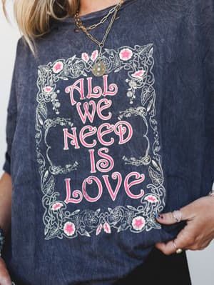 A close-up of a washed charcoal graphic tee reading 'All We Need Is Love' in bold pink lettering, framed by a vintage-style botanical illustration — styled with layered gold chain necklaces and stacked rings for that effortlessly collected, free-spir