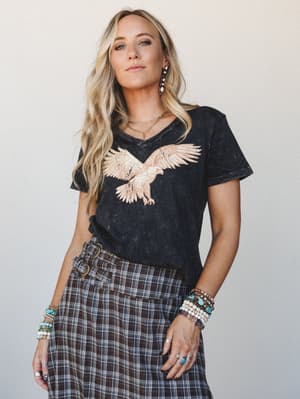 A washed charcoal graphic tee with a rose gold eagle print is tucked loosely into a dark plaid skirt, layered with stacks of turquoise and beaded bracelets — the kind of outfit that feels effortless but clearly has a point of view.