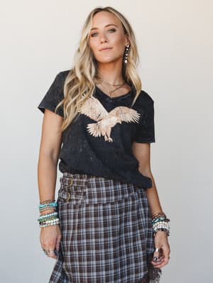 A mineral-washed charcoal graphic tee with a rose gold eagle embroidery paired with a plaid wrap skirt and stacked beaded bracelets — the kind of outfit that looks effortless but clearly took some thought.