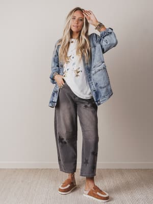 Woman wearing a denim jacket, white shirt with graphics, gray pants, and brown shoes on a plain background