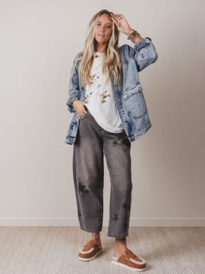 Woman wearing a denim jacket, white shirt with graphics, gray pants, and brown shoes on a plain background