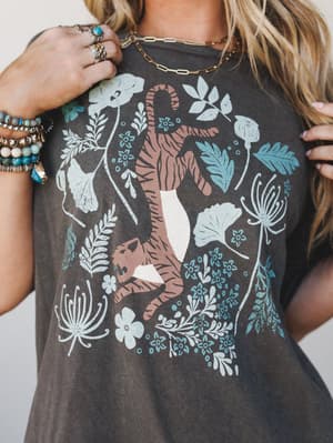 A close-up of a washed charcoal graphic tee featuring a boho tiger-and-botanicals print, styled with layered gold chain necklaces, stacked turquoise and beaded bracelets, and silver rings — the kind of tee that does all the personality work for you.