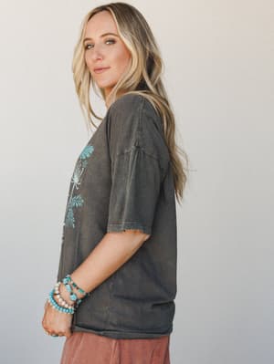 A side-profile shot of a model in an oversized washed charcoal graphic tee with turquoise botanical print details, styled with dusty rose bottoms and a stack of turquoise and pearl beaded bracelets — effortlessly lived-in and boho through and through