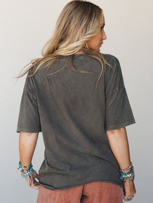 A back-view shot of a model in an oversized acid-washed charcoal tee styled with stacked turquoise and beaded bracelets — effortlessly lived-in and very much the vibe of someone who has great taste without trying too hard.