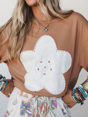 A relaxed mocha tee with a large crochet flower appliqué at center front, styled with a floral print skirt, stacked bracelets, and a turquoise charm necklace — the kind of outfit that looks effortless but clearly wasn't an accident.