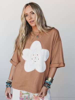 An oversized mocha tee with a large crochet flower appliqué at center front, styled with stacked boho bracelets and a peek of floral pants — casual and expressive in the best way.