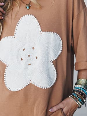 A close-up shot of a mocha oversized top with a large crochet flower appliqué at the center, styled with stacked boho bracelets and a turquoise pendant necklace — the kind of top that does all the talking.