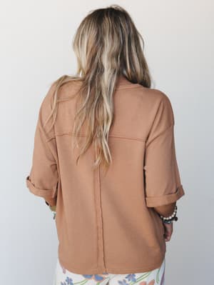 A back view of a relaxed mocha pullover top with rolled sleeves and a subtle center seam detail, paired with floral-print pants and stacked bracelets — effortless and lived-in in the best way.