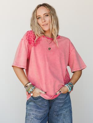 An oversized washed pink tee with crochet floral appliqué at the shoulder and hem, styled with wide-leg cargo jeans and a stack of mixed bracelets — the kind of effortless, personality-forward look that makes a simple top feel like a whole outfit.