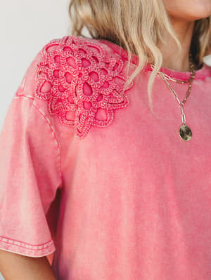 A close-up detail shot of a washed pink-red tee with a standout crochet floral appliqué at the shoulder — the kind of top that makes a basic outfit feel intentional, layered with delicate gold chain necklaces for an easy, lived-in boho look.