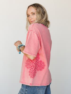 A washed coral-pink oversized tee with a tonal lace appliqué detail at the hip, styled with denim and a stack of turquoise bracelets — the kind of top that looks like you tried just enough.