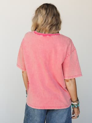 Shot from the back, a model wears an oversized acid-washed coral-red tee with a relaxed drop shoulder and contrast rib neckline, paired with wide-leg denim and stacked boho bracelets — the kind of top that looks like it's been loved for years.