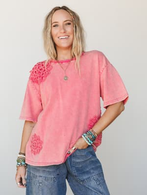 A washed coral-pink oversized tee with floral crochet appliqués at the shoulder and hem, styled with wide-leg denim and stacked bracelets — the kind of top that does all the personality work for you.