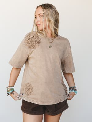 A relaxed, slightly oversized mineral-washed tee in warm mocha, detailed with tonal crochet medallion appliqués — styled with dark chocolate shorts and layers of beaded bracelets for an effortlessly earthy boho look.