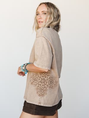 A model shot from behind-ish angle showing a washed mocha oversized top with a stunning crochet lace appliqué across the back hem, paired with dark chocolate shorts and a stack of turquoise beaded bracelets — the kind of piece that makes people ask w