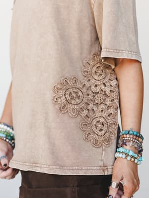 A close-up detail shot of a washed mocha tee with a standout crochet floral appliqué at the hip, styled with layers of beaded and stone bracelets that feel like they've been collected over years of good trips.