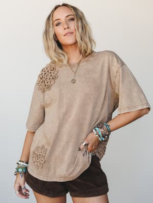 An oversized mineral-washed tee in warm mocha, dressed up with crochet medallion appliqués at the shoulder and hem — styled with dark chocolate shorts and an armful of turquoise and beaded bracelets for that effortlessly collected boho feel.