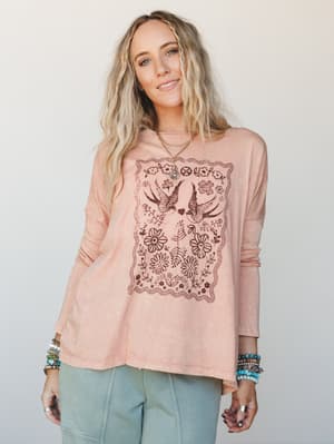 A relaxed long sleeve graphic tee in washed dusty rose, printed with a vintage-inspired bird and floral motif, styled with sage wide-leg pants and layered boho jewelry for that effortless, emotionally attached kind of everyday look.