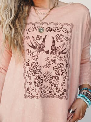 A dusty rose long-sleeve graphic tee with a worn-in, vintage-feel bird and floral print — styled with layered gold necklaces, stacked turquoise bracelets, and easy waves that make the whole thing feel like a Saturday you'd actually want to repeat.