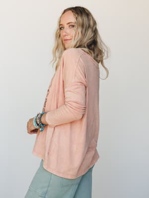 A relaxed long sleeve tee in washed rose, shot from the side to hint at front graphic detailing — styled with sage jeans and a stack of turquoise and stone bracelets for that effortlessly collected, everyday boho feel.