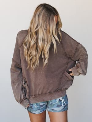 Shot from behind, she's wearing an oversized acid-wash sweatshirt in deep cacao brown with exposed seam detailing, paired with blue camo shorts — the kind of effortless combo that looks like it was zero thought and all instinct.