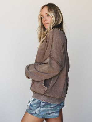 A model shot in three-quarter profile wearing an oversized acid-wash sweatshirt in warm cocoa brown with visible raw seam detailing, paired with blue camo shorts — the whole look reads like a really good Saturday with nowhere to be.