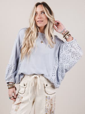 Woman wearing a light blue sweater with bell sleeves and white pants against a beige background