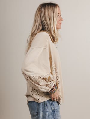 Woman wearing a beige blouse with lace details and blue jeans against a plain background