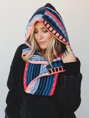 A close, intimate shot of a woman pulling up a bold serape-striped hood on a black velvet pullover — the kind of cozy you actually want to live in, accessorized with layered rings and a delicate necklace.