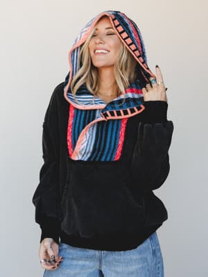 A smiling model pulls up the oversized hood of a black hoodie with a bold multicolor woven stripe panel at the chest and hood lining, styled with light wash jeans and layered boho rings — the kind of top that looks like it came back from a good road