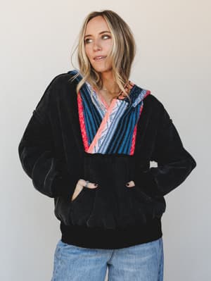 A relaxed, lived-in pullover hoodie in black velvet-textured fabric with a bold woven serape-style panel at the chest and hood, styled with light-wash jeans and layered gold necklaces — the kind of top that feels like a personality, not just a layer.