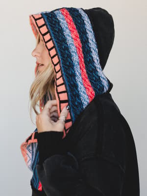 A profile close-up of a model pulling the hood of a black velvet pullover hoodie close, the hood lined in a bold multicolor fair isle knit that does all the talking.