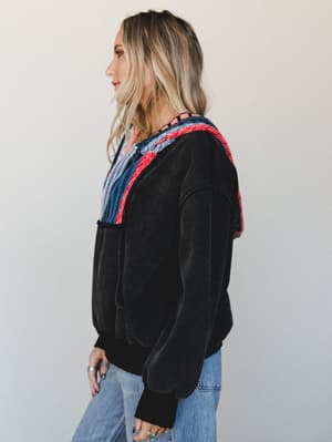 A black velvet-feel pullover hoodie with bold multicolor woven trim along the neckline and shoulders, styled with light wash jeans for an effortlessly cool, lived-in look.
