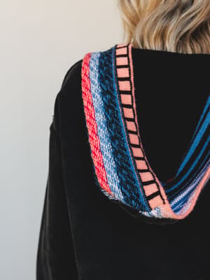 A close-up detail shot of a black pullover hoodie, showing the back neckline and hood edge trimmed with a layered multicolor knit band — coral, red, periwinkle, navy, and black stripes give it an unexpected, artsy-boho feel against the soft black fab