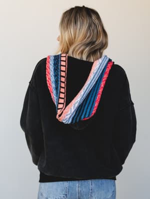 Shot from behind, this black pullover hoodie lets the real star shine — a bold serape-stripe hood lining in red, blue, and blush that makes the whole look feel like a well-traveled weekend.