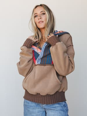 A relaxed, lived-in hoodie pullover in warm mocha with a woven serape-style lined hood, styled with blue jeans and layered rings for that effortlessly personal, cozy-every-day kind of energy.