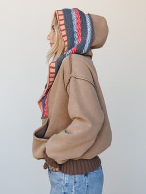 A side-profile shot of a model wearing an oversized mocha hoodie with a bold woven stripe lining inside the hood — the kind of piece that looks like it has a secret personality the moment the hood goes up.