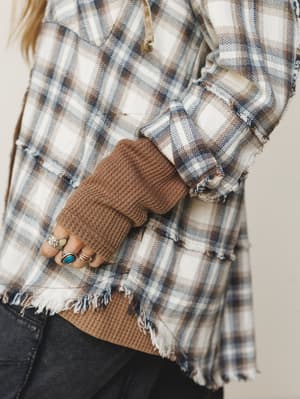 A close-up detail shot of a frayed-hem plaid flannel jacket layered over a camel waffle-knit top, with a hand stacked in silver and turquoise rings — the kind of effortless fall layering that looks thrown together but very much wasn't.