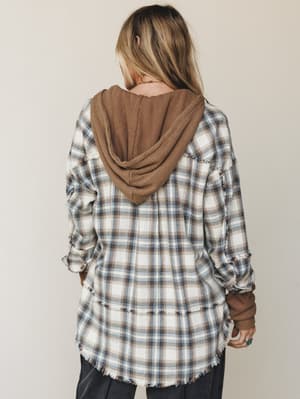 A back-facing shot of a model wearing an oversized plaid flannel jacket with raw fringe edges and a cozy camel waffle-knit hood peeking out — the kind of layered, lived-in look that feels equally at home on a weekend hike or a coffee run.