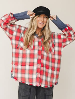 A smiling blonde model adjusts a black newsboy cap while wearing an oversized red plaid flannel hoodie jacket with frayed hem detailing, layered over dark cargo pants — the whole look has that effortless, bundled-up energy of a crisp fall weekend.
