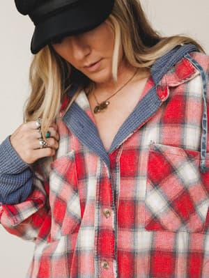 A close-up lifestyle shot of a woman in a red and navy plaid flannel jacket with a blue waffle-knit hood peeking out, layered with stacked silver rings, a turquoise stone ring, and gold charm necklaces — the whole thing reads like a Saturday that sta