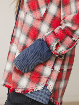 A close-up detail shot of a red plaid flannel jacket with frayed hem trim, layered over a navy waffle-knit top — the kind of effortless, cozy layering that looks like it happened naturally but somehow works perfectly.