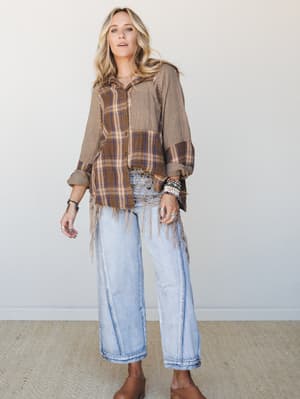 An oversized mixed-plaid button-up with fringe hem trim is styled with light wash wide-leg jeans and stacked boho jewelry — the kind of outfit that looks like it came together effortlessly but absolutely did not.