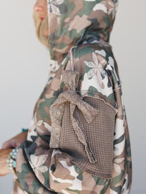A close-up detail shot of a camo hoodie jacket with a mixed-media waffle-knit sleeve panel, tied with a textured bow — the kind of piece that looks like it wandered in from the coolest corner of your closet.