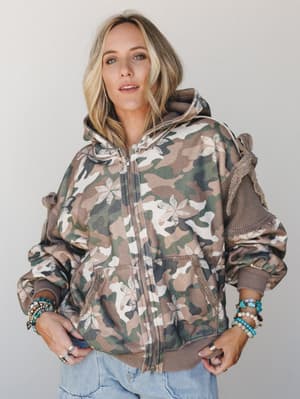 A camo zip-up hoodie jacket with an unexpected floral overlay print gets styled with light wash jeans and stacked beaded bracelets — the kind of piece that feels effortlessly collected, like it was thrifted but actually perfect.